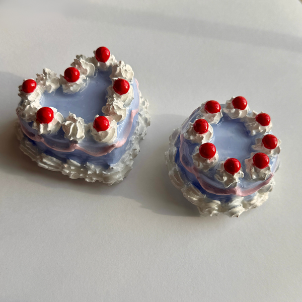 Two small cakes with white frosting, red cherries, and a blue base on a white background.